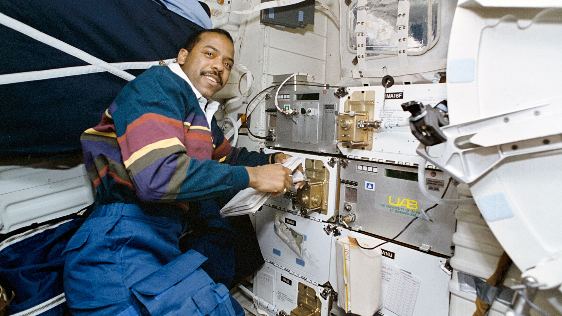 a Black man wearing a rugby shirt and blue pants works on an experiment aboard a space shuttle