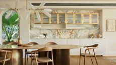 a white kitchen with reeded glass upper cabinets with brass frames. A decorative chandelier sits above a brass and marble dining table, and an archway to an outdoor space is shown to the edge of the frame. 