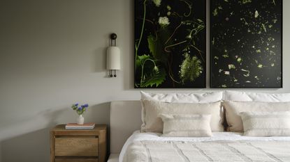 modern bedroom with beige bedding, timber nightstand styled with books and vase, wall sconce, two artworks with black and green, and beige walls
