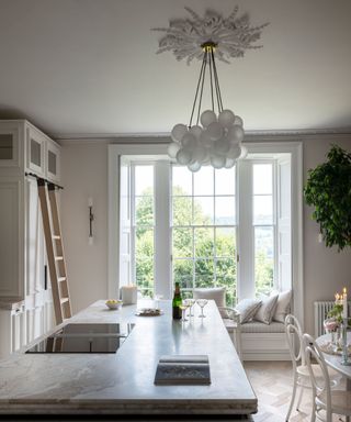 Elegant white kitchen with bay window with window seat and white shutters, wooden flooring, kitchen island with marble countertop, rounded sphere cluster pendant light