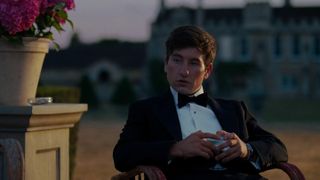 A still from the movie Saltburn of Barry Keoghan as Oliver Quick in a fancy tuxedo sat outside in a chair with a huge looming stately home in the background.