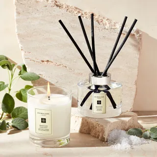 Jo Malone's candle and reed diffuser sit atop a neutrally colored stone slab in a sun-lit environment decorated with greenery and salt.