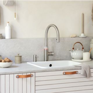 boiling water tap in front of a white sink with a grey splashback and countertop, white cabinets and a grey teapot
