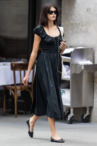 Kaia Gerber walks in New York City wearing a black sundress and black ballet flats