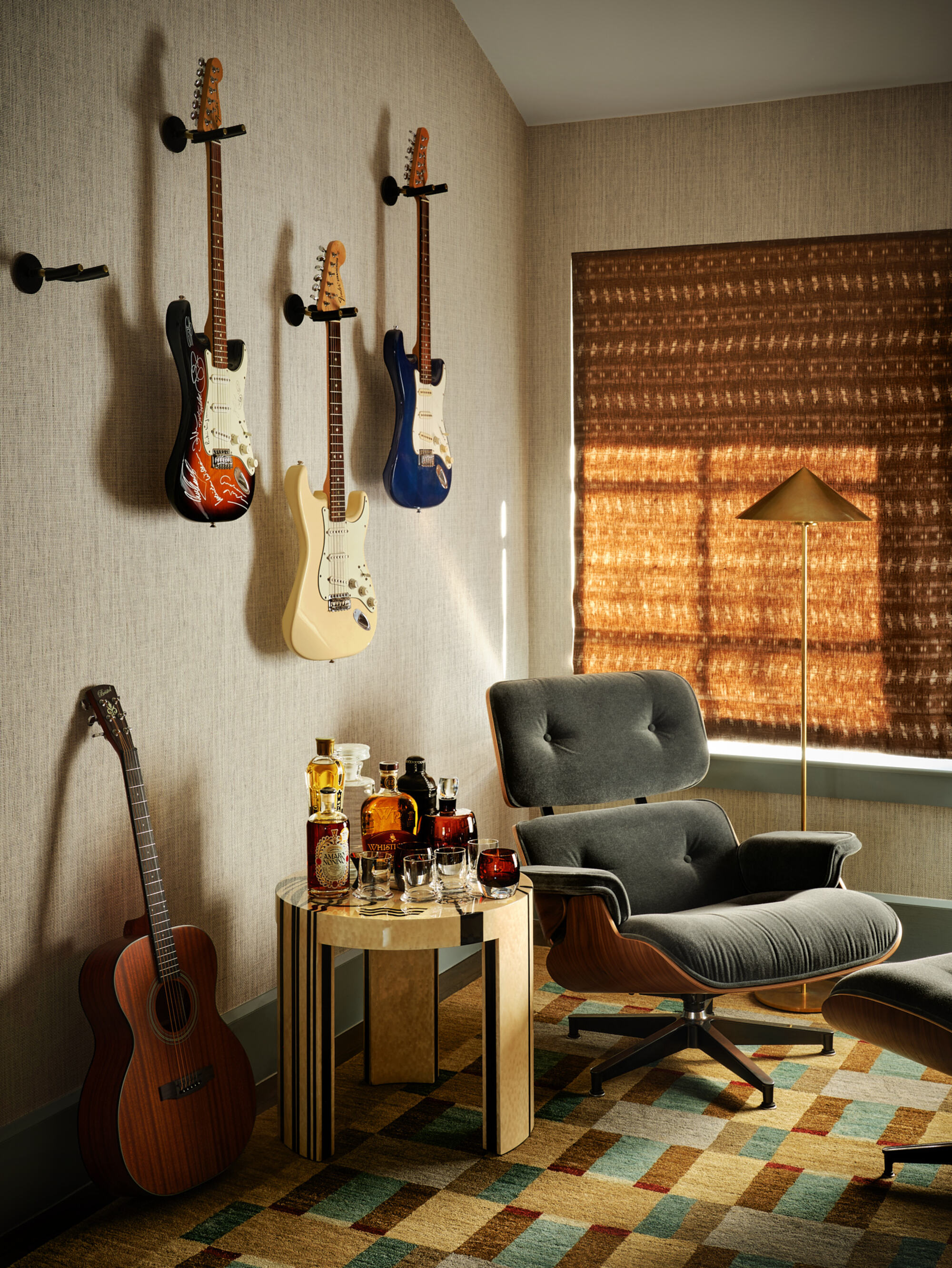 music room with guitars mounted on the wall, an eames armchair, side table styled as a bar, a guitar leaning against the wall, a patterend rug, ochre colored shades, and a gold floor lamp
