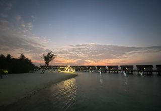 A beach at sunset with an illuminated boat in front of a row of overwater villas in the Maldives.