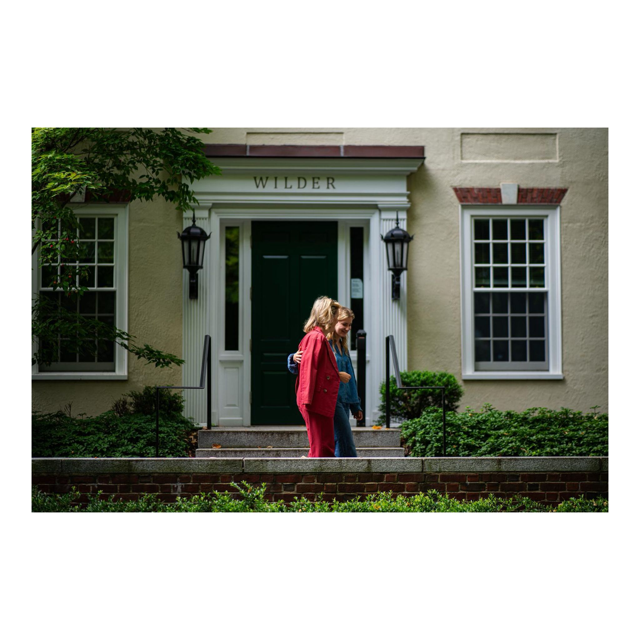 Princess Elisabeth wearing jeans and Queen Mathilde in a pink suit walking past a building that says Wilder
