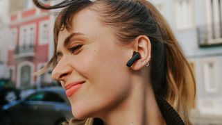 A young woman with blonde/brown hair in a residential street. She has a black Sennheiser Open earbud in her left ear and is smiling, presumably because of music or a podcast
