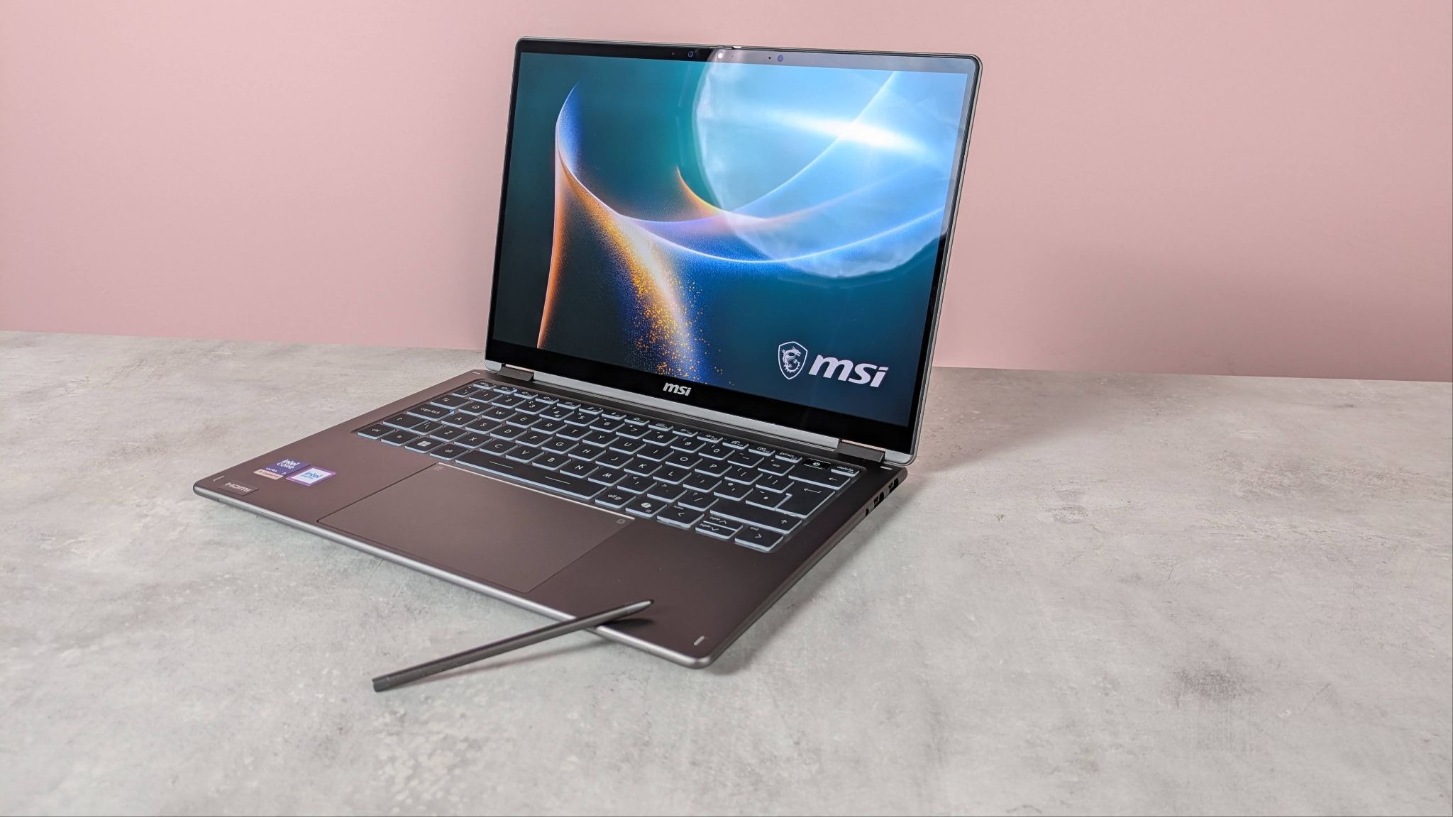 Three-quarter view of MSI Prestige 14 Flip AI+ open on white desk with MSI Nano Pen leaning on it, with pink wall in background