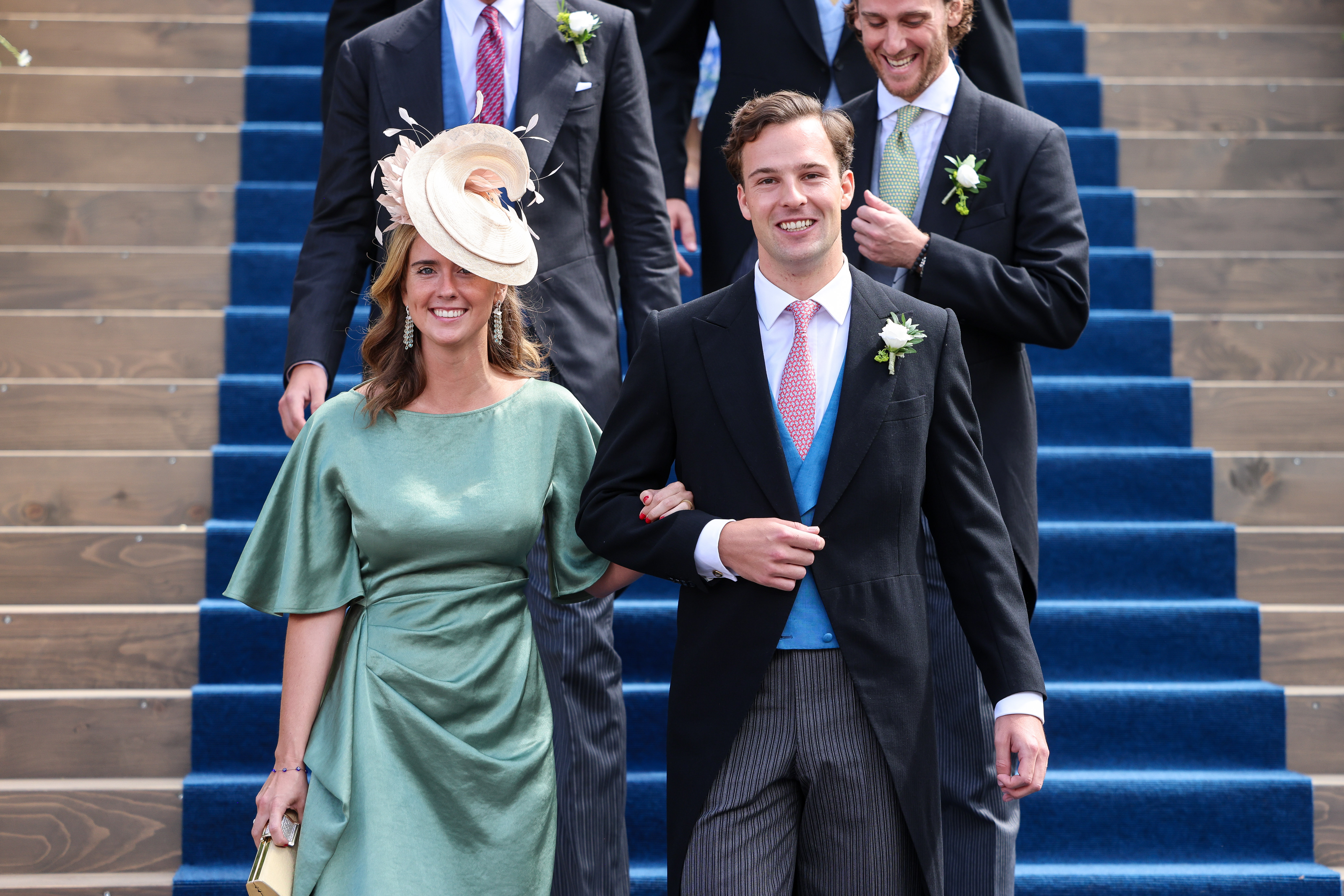 Prince Georg of Liechtenstein wearing a suit and tie smiling walking down a staircase on the arm of a woman in a green dress and hat