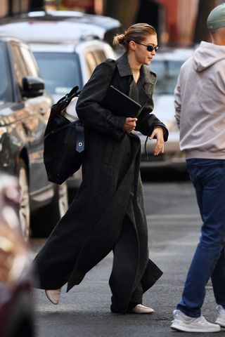 Gigi Hadid steps out in New York, showcasing a dramatic grey wool coat paired with a beige turtleneck and white sneakers. Carrying a black bag and laptop, the model completes the chic look with sleek hair and dark sunglasses.