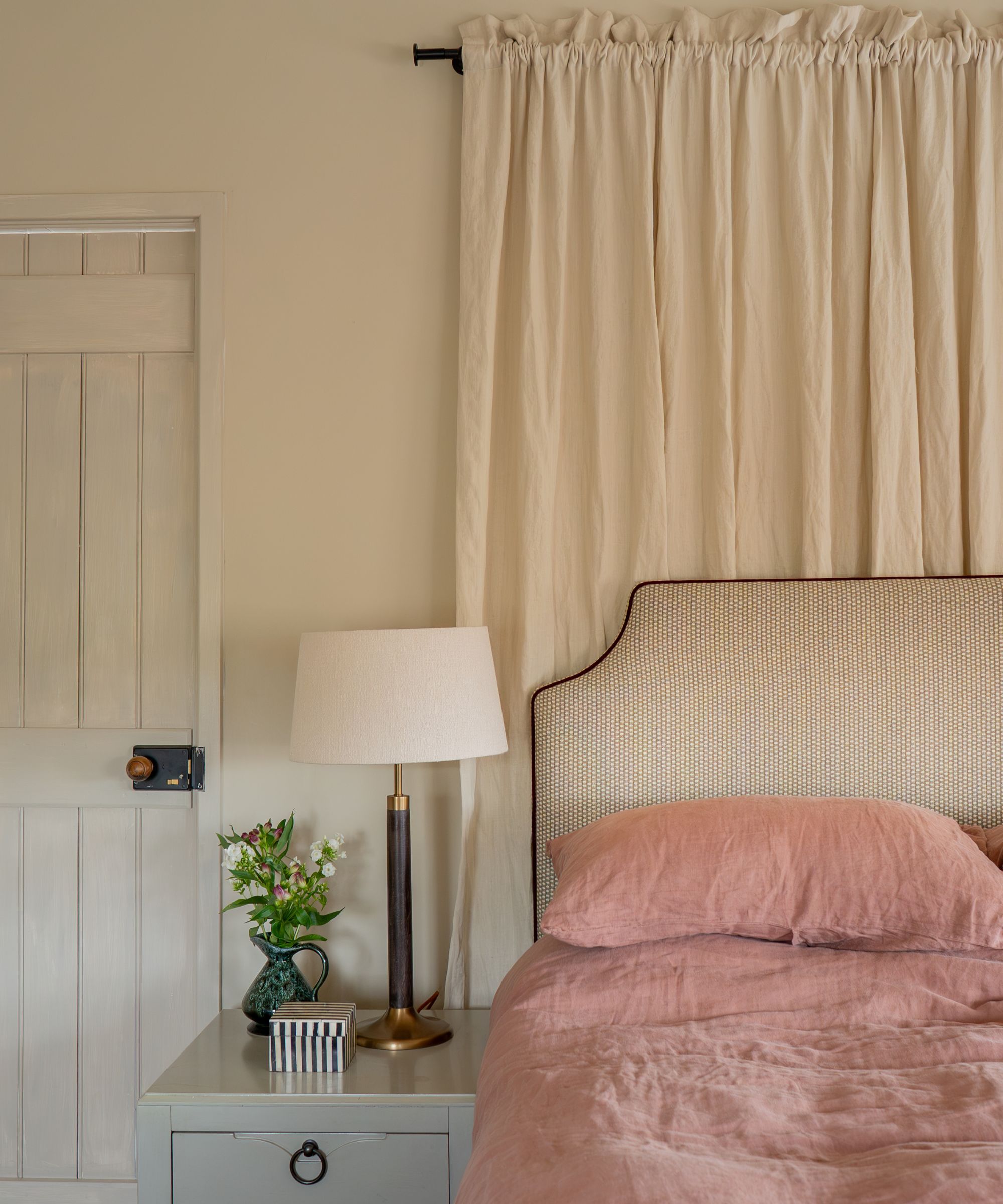 neutral bedroom with a soft neutral canopy above the bed, striped headboard with contrast piping, rose pink bed linen and a small bedside table with a lamp