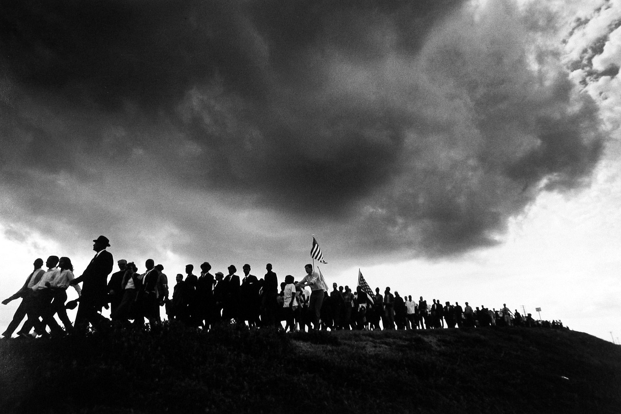 A dense, dark storm cloud looms over a long line of silhouetted marchers carrying American flags as they walk along a ridge during the Selma to Montgomery march.