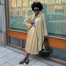 Woman wears trench coat, pleated skirt and grey knit top