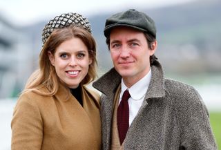 Princess Beatrice, wearing a tan coat and tall headband, standing next to Edoardo Mapelli Mozzi, who is smiling and wearing a gray coat and red tie with a flat cap, standing outside at a racecourse