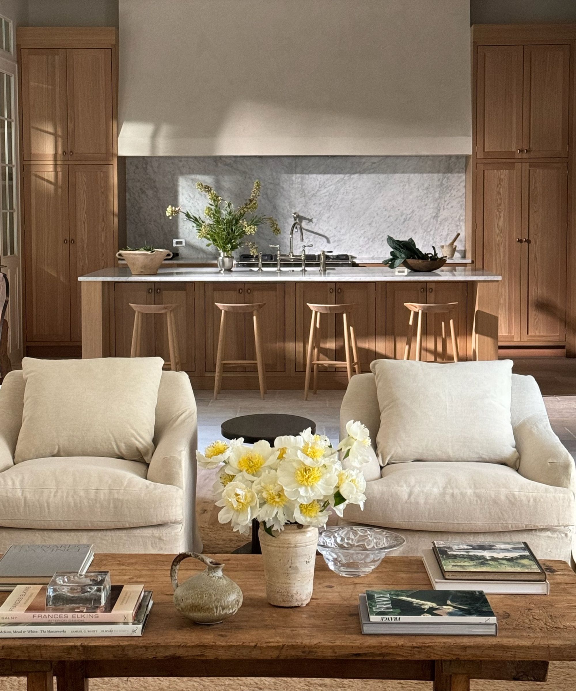 Two cream armchairs sitting behind a rectangular coffee table with flowers and books sitting on top. Open plan kitchen in the background with wooden cabinetry and bar stools.