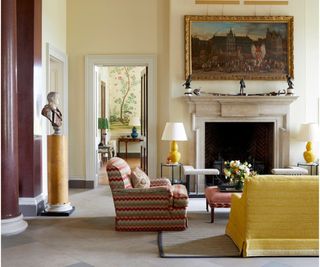 Grand living room with a large fireplace, ottoman, fender, armchair, sofa and two yellow table lamps with small lightweight side tables either side of the fire