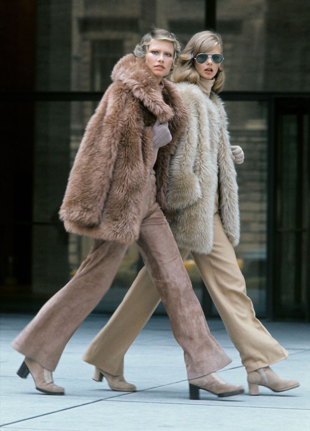 Two models walking in front of the Seagrams Building in New York City wearing furry coats, pants and grey suede boots - left model: beige furry coat by Bill Blass, camel color cashmere pants and beige cashmere turtleneck with Kay Fuchs gloves and Palmroth for Fiorentina boots - right model: cinnamon-russet, fluffy coat by Anne Klein, same color chamois pant and turtleneck with Brousseau gloves and Charles Jourdan boots