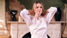 A young woman dressed in chef attire, with blonde hair gathered in a low pony tail, touches the back of her shoulders with both hands while standing in a craftily decorated room with distressed cement green and beige walls, an antique fireplace, sculptures, and ceramics.