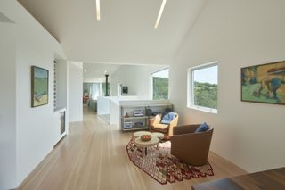 Interior view of a living area at Triple Barn house featuring white walls, wood flooring, two brown armchairs with blue cushions, a curved table with a bowl on top, a multicoloured patterned rug, wall art, windows and a shelving unit with various items