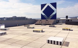 Square-shaped structures are set on the rooftop. Each structure has black & white lines on its side, and the top is in different colors - yellow, blue, and white. In the distance, a wide and tall, square-shaped structure is set, in deep blue, with a rhombus-shaped mirror.