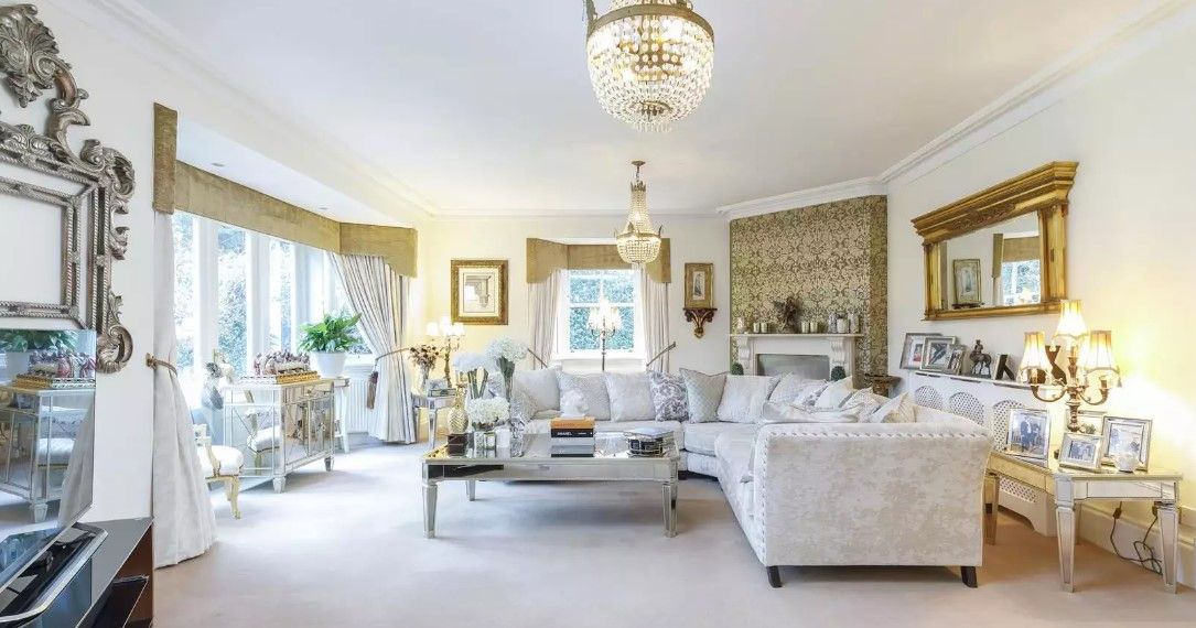 The white living room shows a Victorian mirror on one wall and a chandelier above with a corner piece white sofa and bifold doors which lead onto the patio. There is also a completely white carpet
