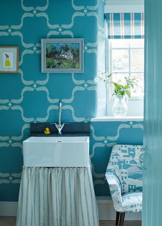 A blue powder room with a white sink, a fabric sink curtain and blue and white patterned wallpaper