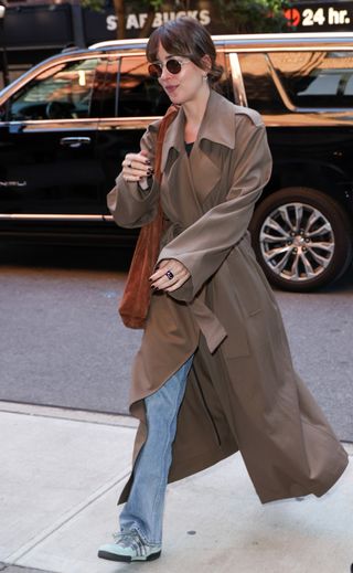 Dakota Johnson wearing a brown trench coat and jeans with a brown suede slouchy bag.