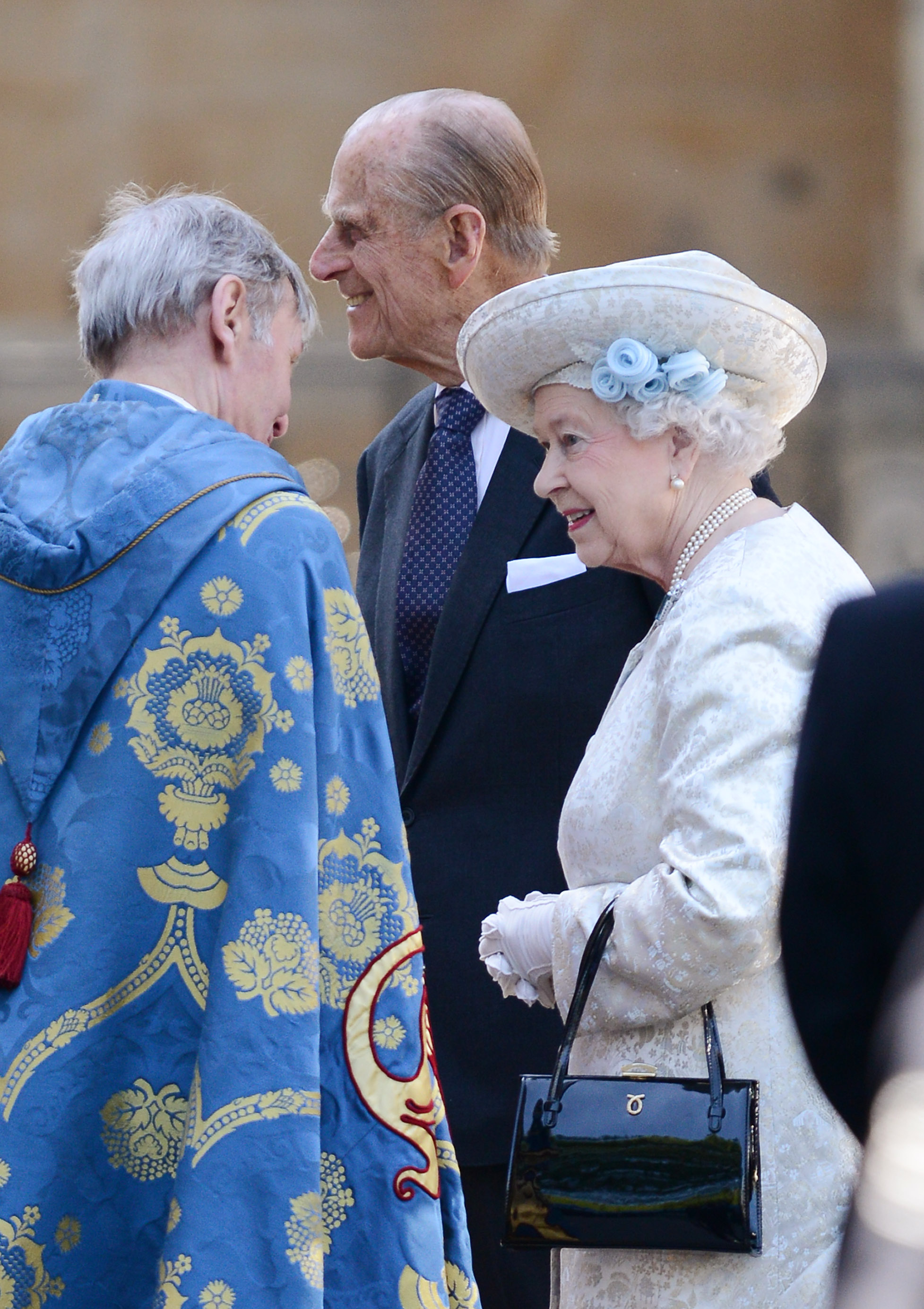 Queen Elizabeth wearing a white coat and hat standing next to Prince Philip talking to a clergyman