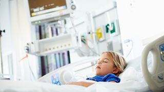 A young girl lies sleeping in a hospital bed with a breathing tube in her nostrils