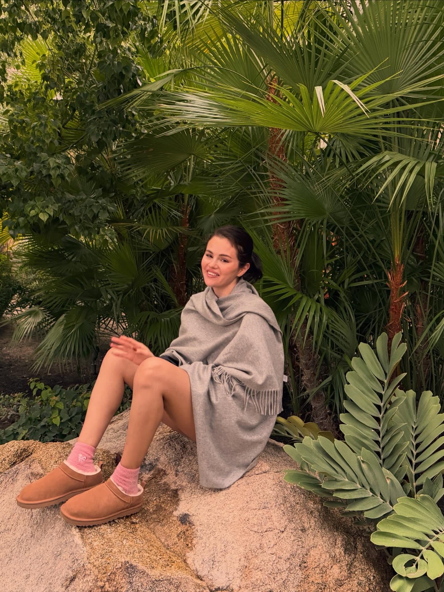 Selena Gomez sits on a large rock surrounded by trees and shrubbery wearing a scarf knit with Uggs and pink socks.