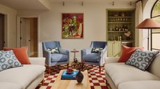 a living room with a checker-printed rug, two blue accent chairs, two white sofas with pillows on top, a green built-in bar on the right side, and abstract artwork on the wall