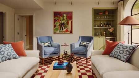 a living room with a checker-printed rug, two blue accent chairs, two white sofas with pillows on top, a green built-in bar on the right side, and abstract artwork on the wall