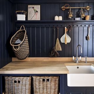 Utility room with navy wall panelling, oak counter and rattan baskets.