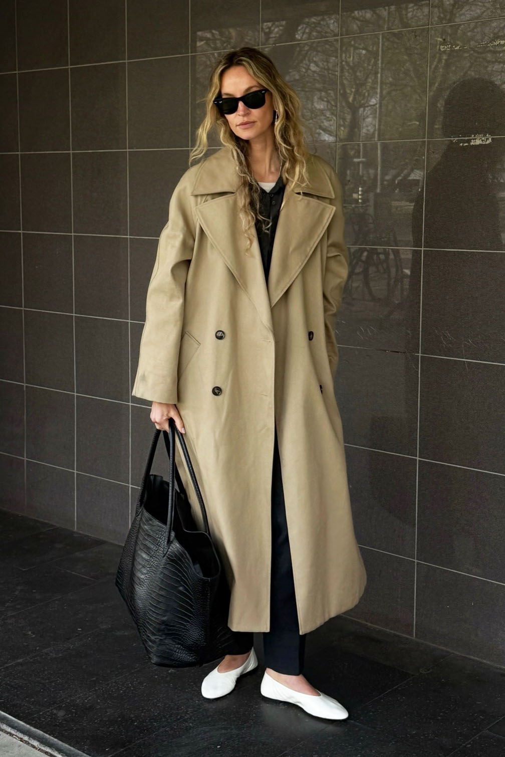 a woman wearing a trench coat, white flats, black sweater, black pants, and leather bag