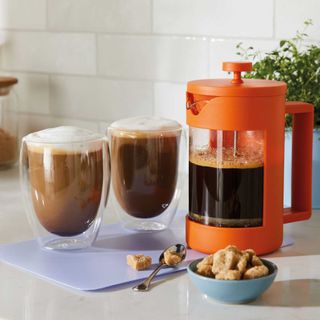 Orange cafetiere with coffee inside, two cups of coffee and brown sugar on kitchen worktop