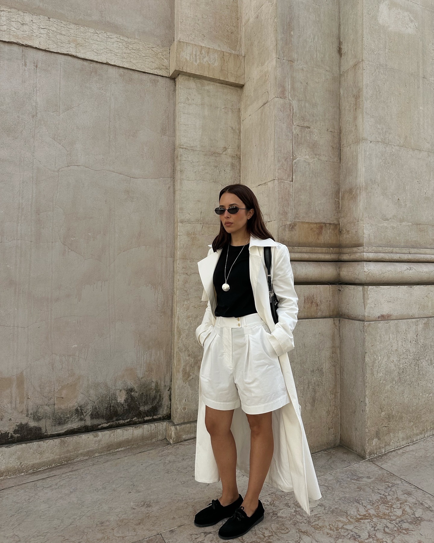 Influencer @deborabrosa wears white linen shorts with a black top, a white trench coat and suede boat shoes.