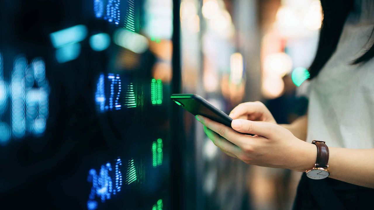 A woman looks at her smartphone in front of a stock-ticker screen