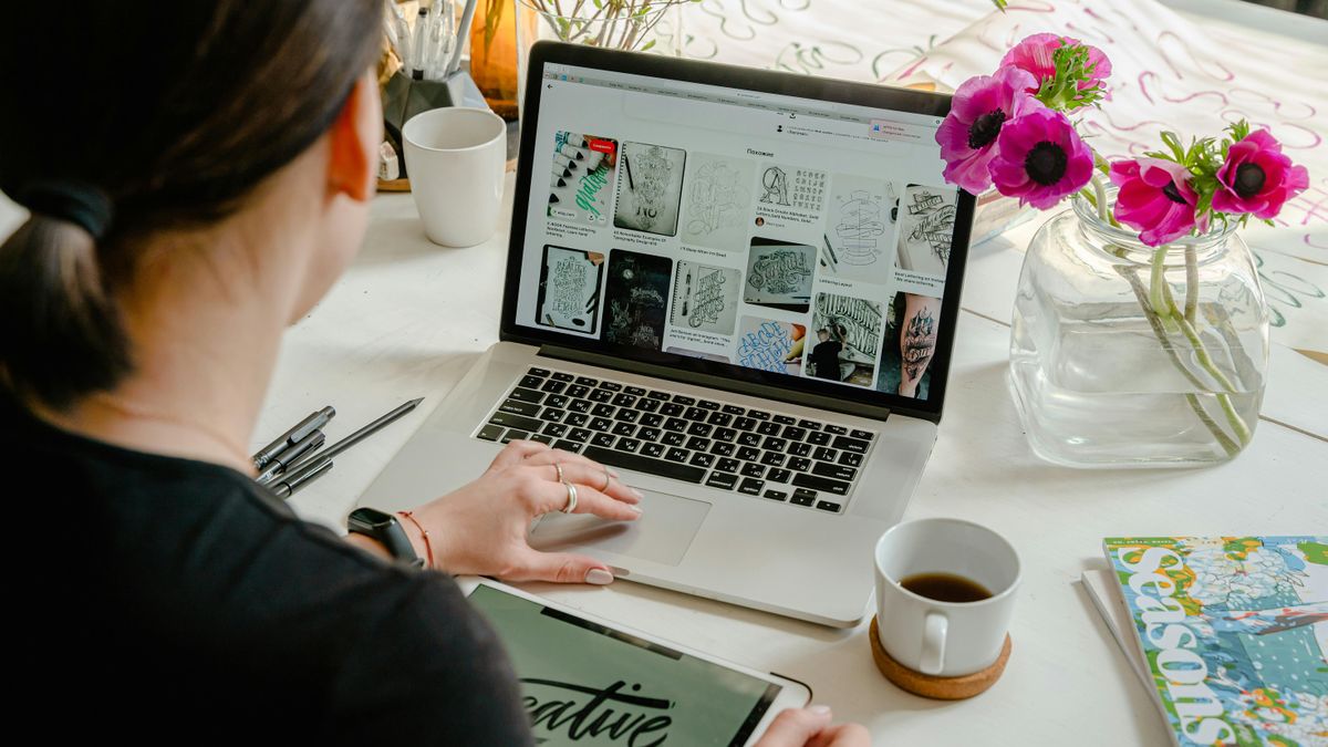 Woman looking at laptop screen, holding iPad and coffee next to her hand