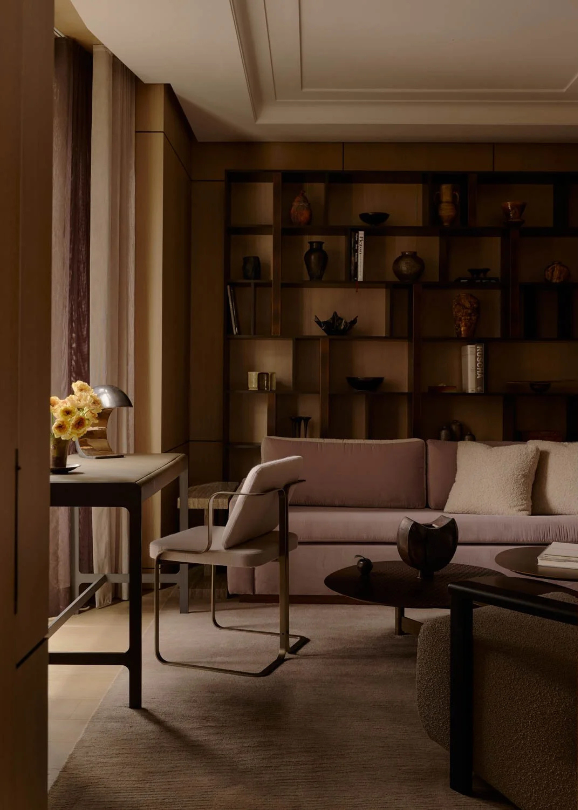 A brown living room with a wall of shelves holding vases and books near a plush couch, a coffee table and a study space with a vase of flowers