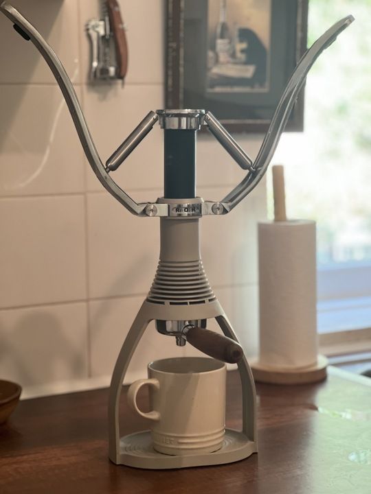 A gray ROK coffee machine with the arms up and a white ceramic mug in the base. The kitchen counters are wooden, and there is a paper towel holder on the counter.
