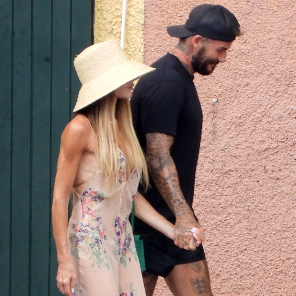 Victoria Beckham walks down the street holding hands with David Beckham wearing a sheer floral maxi dress with brown sandals.