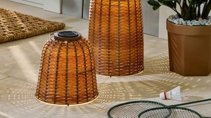 Pair of woven outdoor lanterns styled on a back porch alongside a potted plant and badminton accessories.