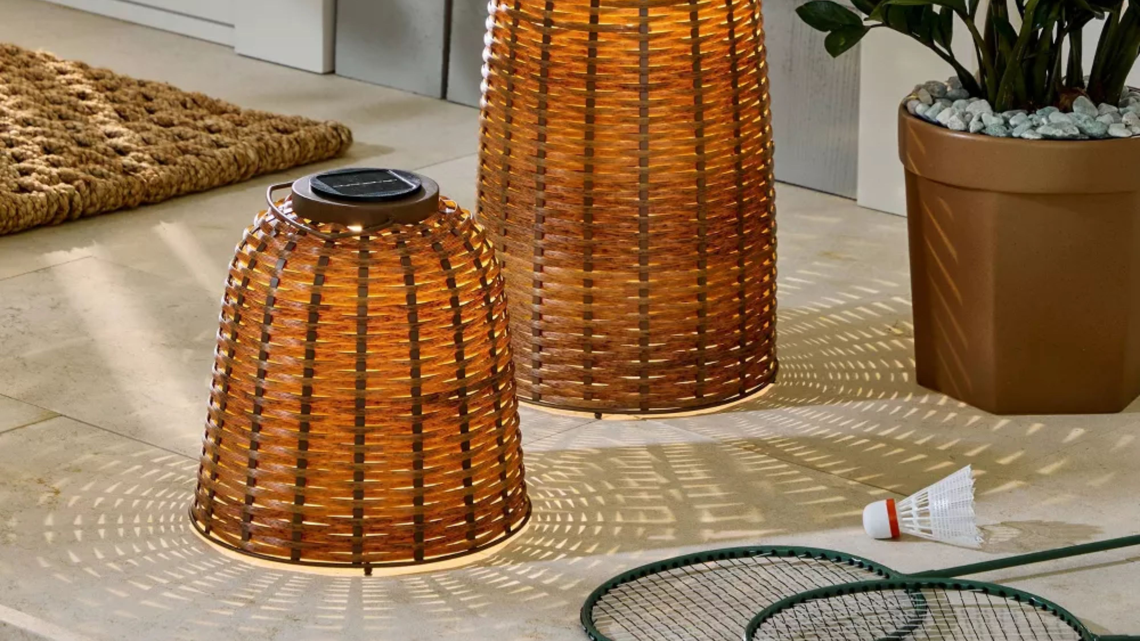 Pair of woven outdoor lanterns styled on a back porch alongside a potted plant and badminton accessories.