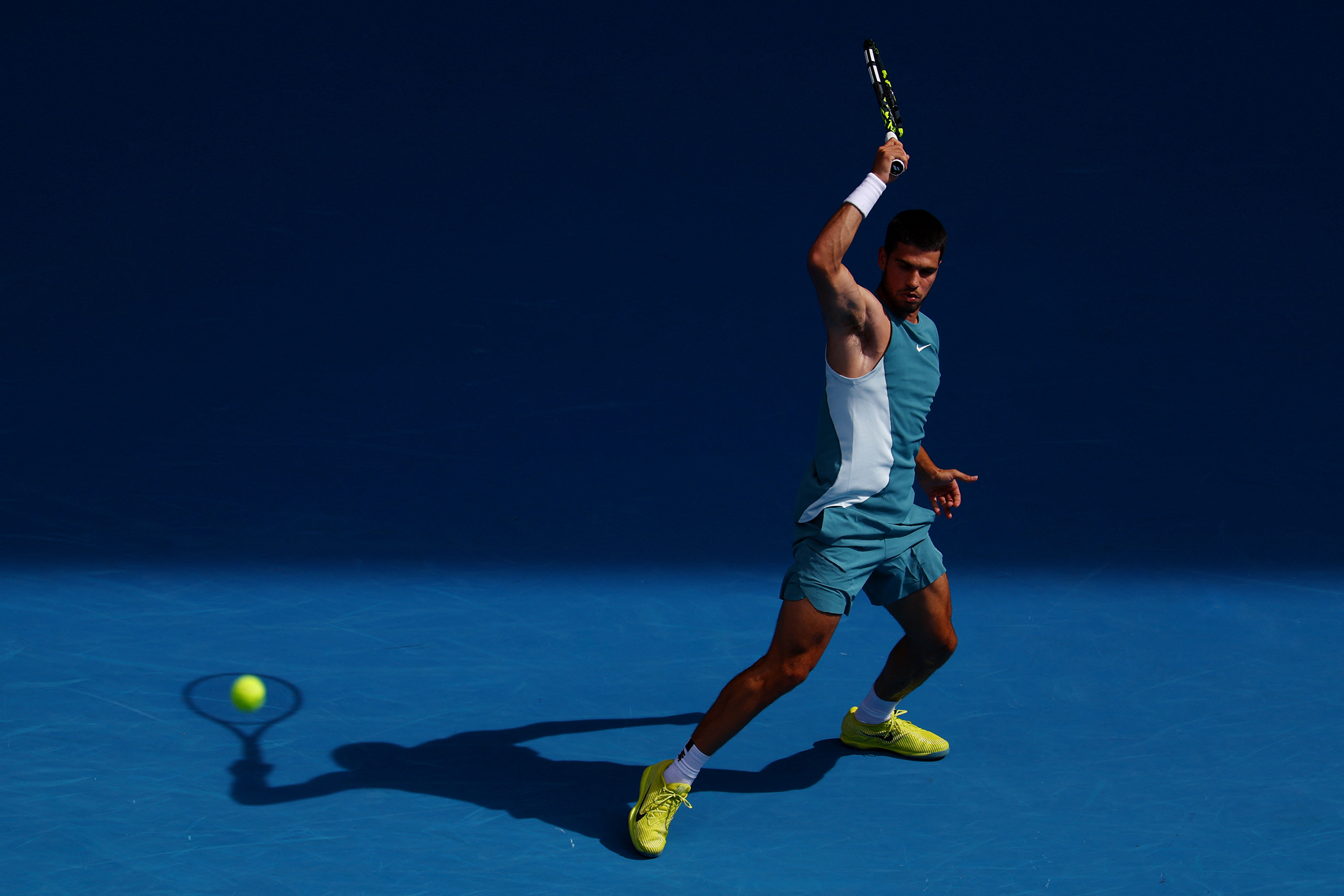 A tennis player in a turquoise outfit prepares to swing a racket on a blue court, with a ball in mid-air. Bright spotlighting creates shadows