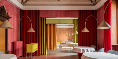A surreally colorful, retro-futuristic restaurant features a red, rose, olive green, and orange palette, punctuated by flowy curtains of each hue, sculptural standing lights in brass, and mid-century trolleys in brass and eye-catching yellow.