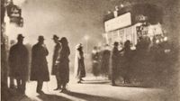 A grainy photogravure of several coated, hatted figures standing in silhouette on a pavement, their long shadows stretching across the ground beneath a glowing street sign.
