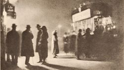 A grainy photogravure of several coated, hatted figures standing in silhouette on a pavement, their long shadows stretching across the ground beneath a glowing