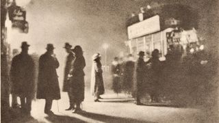 A grainy photogravure of several coated, hatted figures standing in silhouette on a pavement, their long shadows stretching across the ground beneath a glowing street sign.