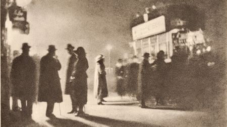 A grainy photogravure of several coated, hatted figures standing in silhouette on a pavement, their long shadows stretching across the ground beneath a glowing street sign.